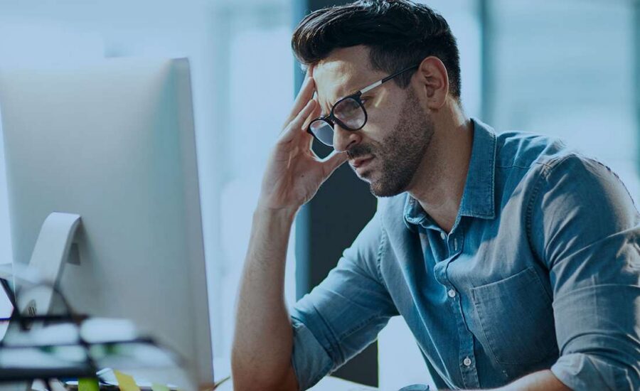 Frustrated man in a denim shirt sits at a desk, hand resting on his forehead while looking at a computer screen.