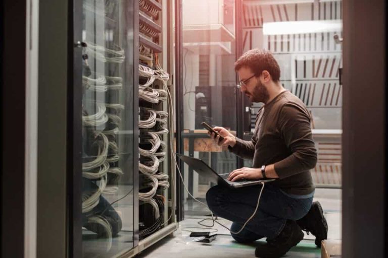Techniker kniet neben Serverracks in einem Rechenzentrum und benutzt einen Laptop und ein Smartphone.